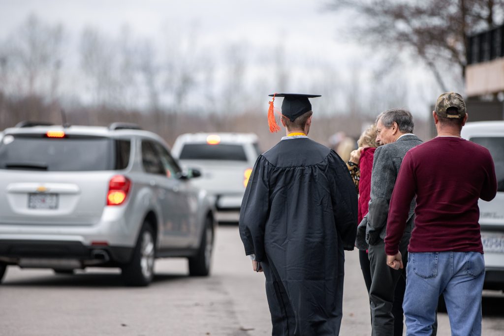 A graduate and others walk away next to a line of cars
