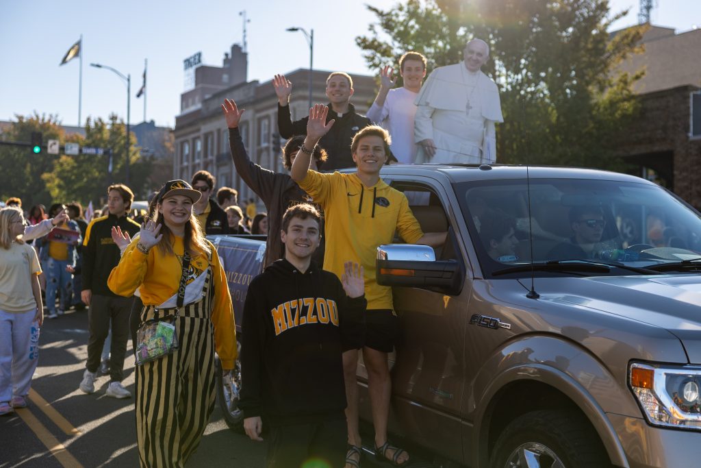 Students in a homecoming parade stand next to a truck and wave